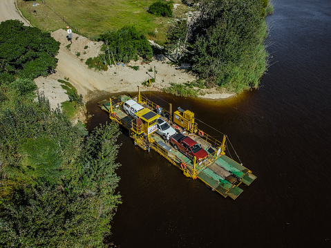 Aerial shot of a moving yellow and green ferry carrying 2 white, and 1 orange vehicle over a brown water river from a sandy beach surrounded by green bushes and trees, Malgas.
