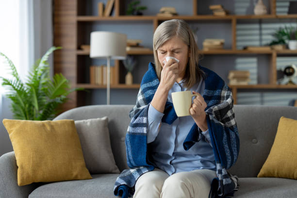 istock Senior woman feeling unwell while resting wrapped in checkered blanket and getting warmed with hot drink in ceramic cup. Sneezing lady blowing nose to paper napkin and antiviral tea indoors 2148444923