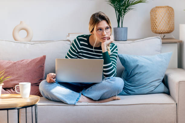 istock Pretty young bored woman working with her laptop while sitting on couch at home. 2148445677