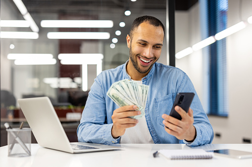 Smiling hispanic young man sitting in a formal office at a table, holding cash money banknotes in his hands, using a mobile phone.