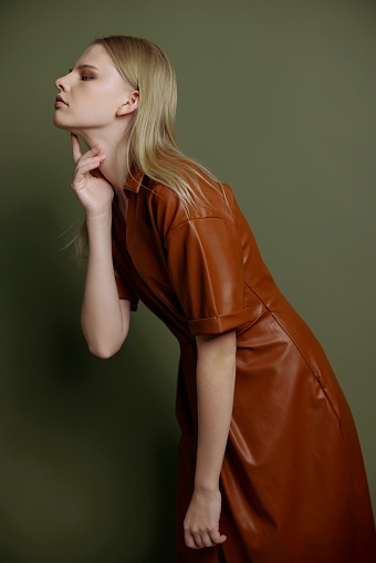 Fashion profile studio portrait of blonde stylish young woman in trendy leather trench coat posing isolated on olive green background. Resale offer for marketplace. Huge sale concept