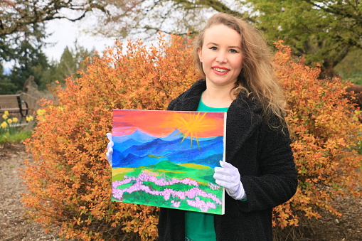 A Russian woman holding her original painting. She is wearing long, red, curly hair, makeup, a black fully unbuttoned coat, a green sweater, white gloves and holding her painting. She is in front of a bush in orange Spring color in a public park.