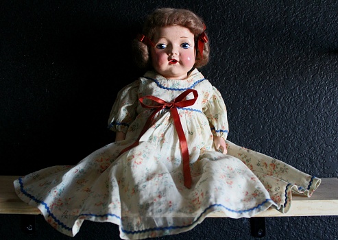 1930's baby doll. Wearing a pretty floral dress with blue trim. Red bow on the dress and in her hair. Red little lips and blue eyes that sparkle.