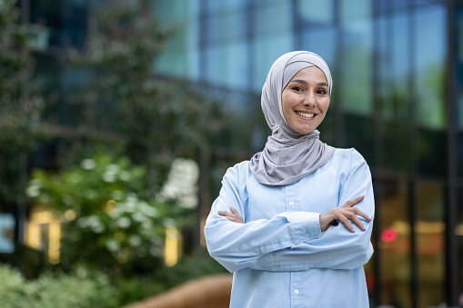 Portrait of a cheerful Muslim woman with hijab, confidently standing with crossed arms in a modern city environment.
