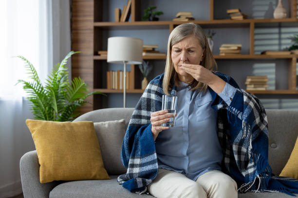 istock Sad elderly lady sitting on pillowed sofa under blanket and getting ready for taking medicine with glass of water. Struggling woman suffering from headache and using painkillers during sick leave 2148448946
