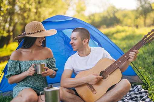 A man plays the guitar while a girl enjoys music, they are camping in nature.