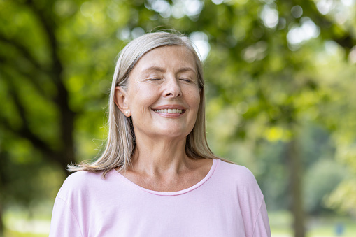 Close-up portrait of smiling senior gray-haired woman in pink t-shirt standing in park and resting, breathing relaxed with eyes closed after walking and doing sports.