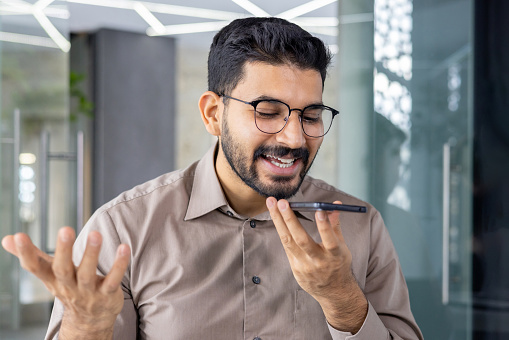 Smiling young adult male engages in a cheerful conversation using a hands-free mobile phone in a modern office setting.