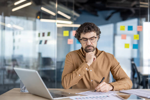 istock Serious thinking businessman behind paper work inside office, man fills forms, reports and invoices. An employee uses a laptop for paperwork 2148453081