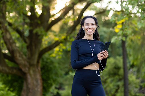 An athletic Hispanic woman smiling and holding a smartphone with earphones, taking a break from her run in a lush green park.