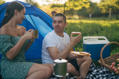 A young man and a woman are camping alone in nature, sitting on a blanket next to the tent and having breakfast.