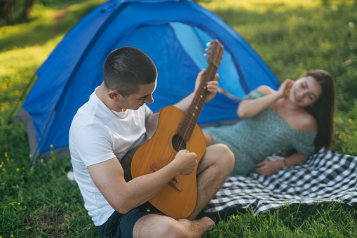 Young adult couple relaxing in nature, sitting next to their tent camp. A girl enjoys listening while her boyfriend plays the guitar.
