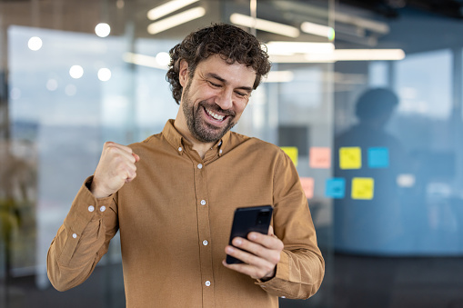 Joyful male entrepreneur excitedly pumps fist in victory as he looks at his smartphone, standing in a modern office setting.