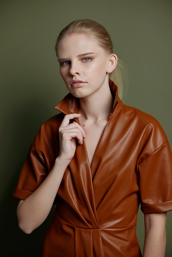 Resale New collection offer. Elegant blond young lady in trendy leather trench coat dress looks with blue eyes at camera presenting evening makeup posing isolated on olive green studio wall background