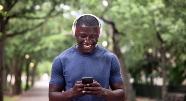 Phone, fitness and happy black man in a park walking with headphones, music or chat in nature. Smartphone, search and male runner in a forest with app for workout tracking, progress or exercise goal