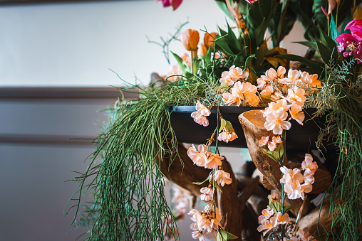 An elegant arrangement of artificial peach flowers on a wooden shelf, surrounded by greenery and hints of orange and pink flowers. Perfect for interior decor.
