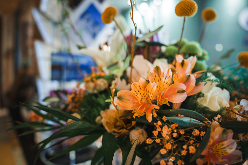 Colorful floral arrangement featuring vibrant orange lilies, small orange flowers, yellow button like blooms, and varied greenery in a blurry indoor setting.