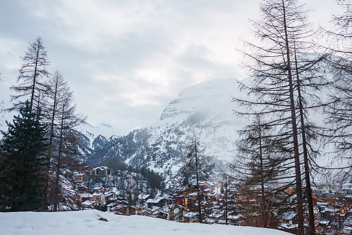 Winter wonderland scene in a mountainous area with snowy village, bare trees, and misty peaks. Serene and tranquil ambiance, possibly in Alps or Dolomites.