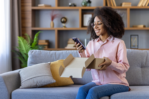 A happy buyer of an online store is sitting on a sofa in the living room of a house at home, a satisfied woman is holding a cardboard box with a purchase of goods in her hands, and a phone.