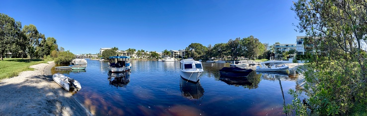 Panoramic view of a variety of boats moored in the calm waters of a canal, native tropical coastal forest, houses and resorts on a sunny day. Noosaville, Noosa Heads, Sunshine Coast, Queensland.