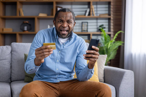 istock African american man sitting on sofa while holding mobile phone and plastic bank card with stressed facial expression. Adult male buyer making unsuccessful attempts paying for online order at home 2148462470