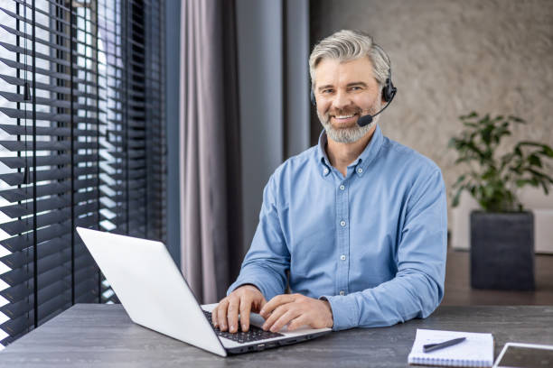 istock Senior grey-haired man in wireless headset sitting in home office with laptop and notebook. Smiling entrepreneur getting ready for online conference and discussing planning of working strategy 2148462891