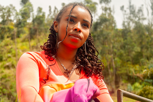 A woman adorned in a vibrant orange blouse radiates a sense of calm as she rests, her gaze reflecting deep thought or gentle reverie. The lush green background suggests a natural and serene environment that invites introspection and peace.