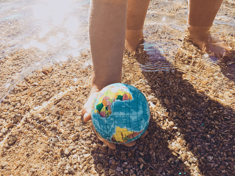 A child plays with a world globe ball by the sea in sunshine. Taken from a personal perspective viewpoint. The image is taken with a mobile phone for a modern style. This is a mass produced world sponge ball from a dollar shop, the map has been blurred. CONCEPT - Child with the world in his hands.