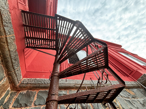 A spiral staircase is seen from the side of a building. The staircase is made of metal and is rusted. The building is red and has a stone facade. The sky is cloudy
