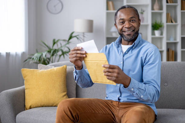 istock Satisfied african american male opening yellow letter envelope while sitting on couch at comfortable living room. Glad adult man in casual outfit receiving great news from relatives via postage 2148465529