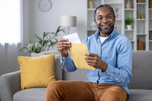 Satisfied african american male opening yellow letter envelope while sitting on couch at comfortable living room. Glad adult man in casual outfit receiving great news from relatives via postage.