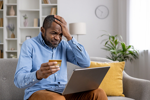 Frustrated cheated man received rejected transfer , error of buying products in online store, african american mature holding bank credit card in hands, using laptop sitting on sofa in living room.