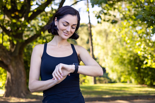 Smiling woman wearing a fitness tracker checks progress during an outdoor exercise session amid greenery.