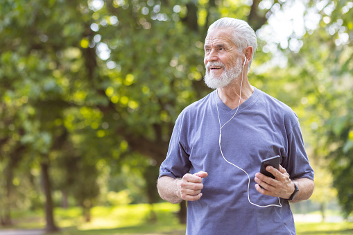 Gray-haired senior man doing sports and running in park, holding phone in headphones, listening to music and smiling.