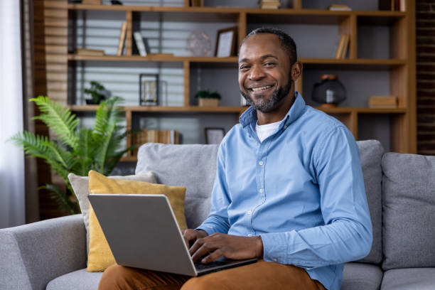 istock Smiling african american man using laptop on couch in stylish home interior 2148468430