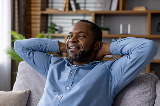 A content African American male sitting on a sofa at home, hands behind head, with a look of peaceful relaxation.