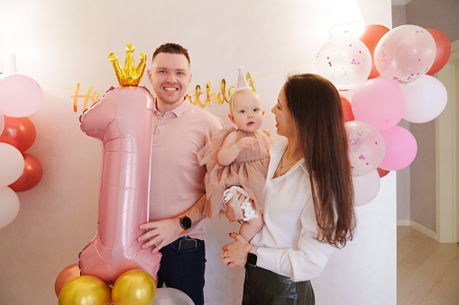 Happy and gentle parents hold baby daughter in pink dress in their arms with birthday balloon decor. Mom and Dad's love. First Birthday House Party.