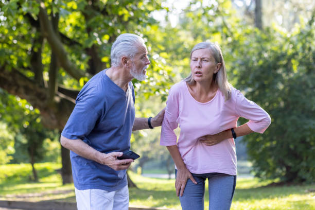 senior couple doing sports and walking in the park, the woman feels severe pain in the side of the body and in the heart, the man tries to help, calls an ambulance on the phone - kalp-krizi stok fotoğraflar ve resimler