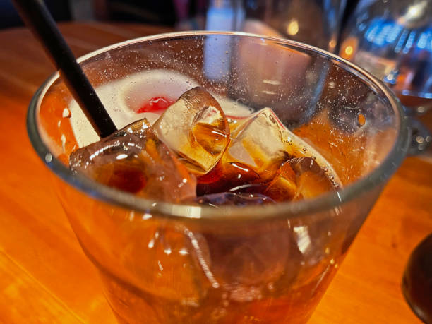 Large glass of soda with ice cubes.    A black straw on left.   On a table.