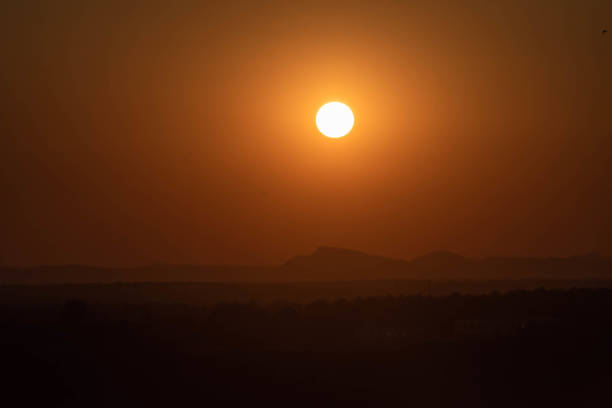 The sun casts a warm orange glow as it sets over a peaceful desert, silhouetting the distant mountains against a hazy sky.