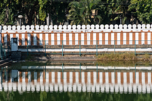 A tranquil waterbody at Shravanabelagola is captured reflecting the pattern of an orange and white wall under clear skies.