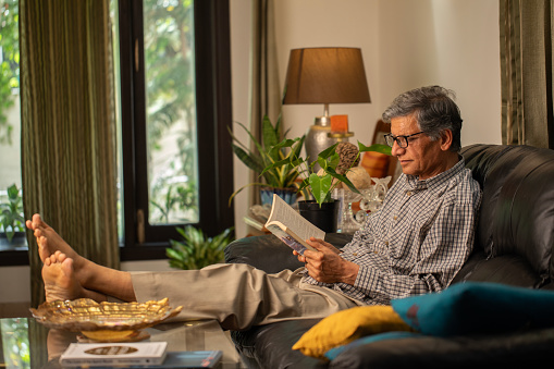 Serious old man in eyeglasses reading book with focus while relaxing on sofa in living room at home
