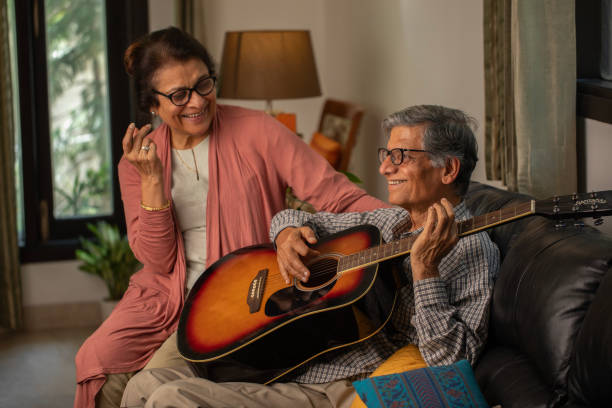istock Elderly wife looking at cheerful senior husband singing and playing guitar while sitting on sofa in living room 2148475654