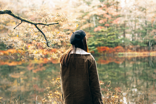 Inspiration, contemplation, freedom, loneliness concept. Back view of young woman in coat standing on shore by water and looking away at lake, outdoors.