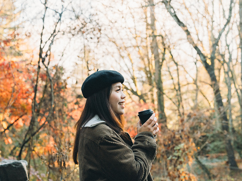 Captured in this autumnal photograph, a young woman stands outdoors, embracing a moment of tranquility and comfort. With a black beret atop her head and a cozy brown sweater wrapped around her, she holds a steaming cup of hot beverage in her hands, smiling as she takes in the scenery. The backdrop of autumn trees, displaying a vibrant palette of greens, oranges, and reds, adds depth and texture to the scene. This image captures a tender moment of travel, evoking a sense of peace and warmth that is inherent in autumn. Whether wandering through the streets on a crisp autumn day or immersing oneself in the natural beauty of the season, travel always holds the promise of unexpected joys and touching moments.