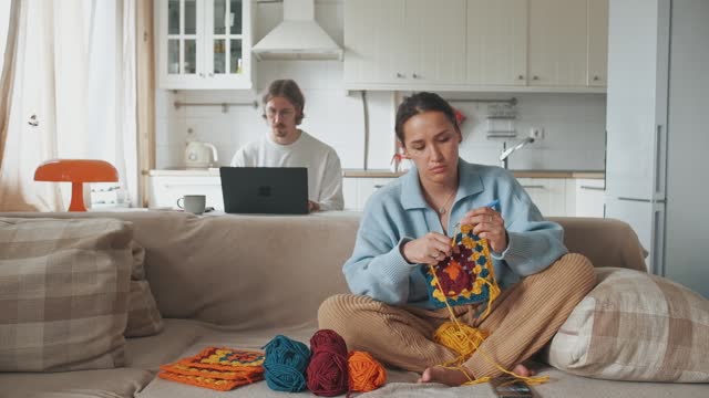 Young woman knitting crocheting with colored yarn granny square while watching online tutorial on phone. And young man working on laptop from homeWoman watching needlework lessons, home hobbies