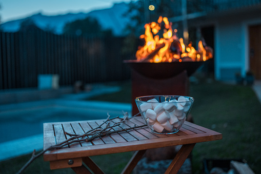 A twilight scene displaying a bowl of marshmallows ready for toasting beside a roaring backyard fire pit, with a serene mountain backdrop.