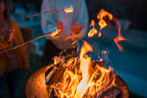 A lively group enjoys toasting marshmallows over a blazing bonfire, clad in casual attire with skewers in hand, at an outdoor gathering.