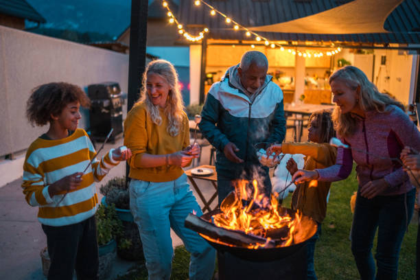 A diverse group engages in lively conversation while roasting marshmallows over an open fire, with joyful expressions and casual evening attire, in a backyard setting.