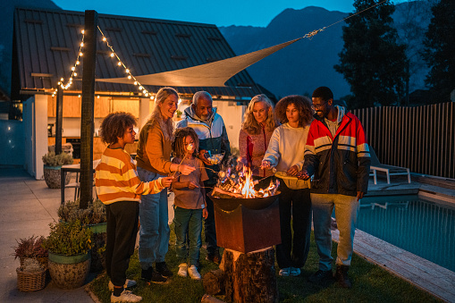 A diverse group gathers around a fire pit at dusk, engaged in roasting marshmallows. The joyful family dons casual attire, connecting across generations in a cozy backyard setting.